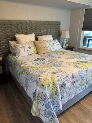 A neatly made bed with floral bedding, a headboard with a wavy design, and a bedside table with a lamp and a plant. - Reel’em Inn - a Waterfront Home on the Canal (Aransas Pass)