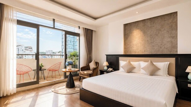 Une chambre d’hôtel dotée d’un grand lit, offrant une vue sur la ville grâce à de larges fenêtres du sol au plafond, d’une table ronde avec des chaises et d’une tête de lit moderne.