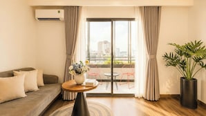 Une chambre d’hôtel moderne avec un canapé, une table ronde agrémentée d’un vase de fleurs, une plante en pot et une vue sur la ville grâce à de grandes fenêtres.