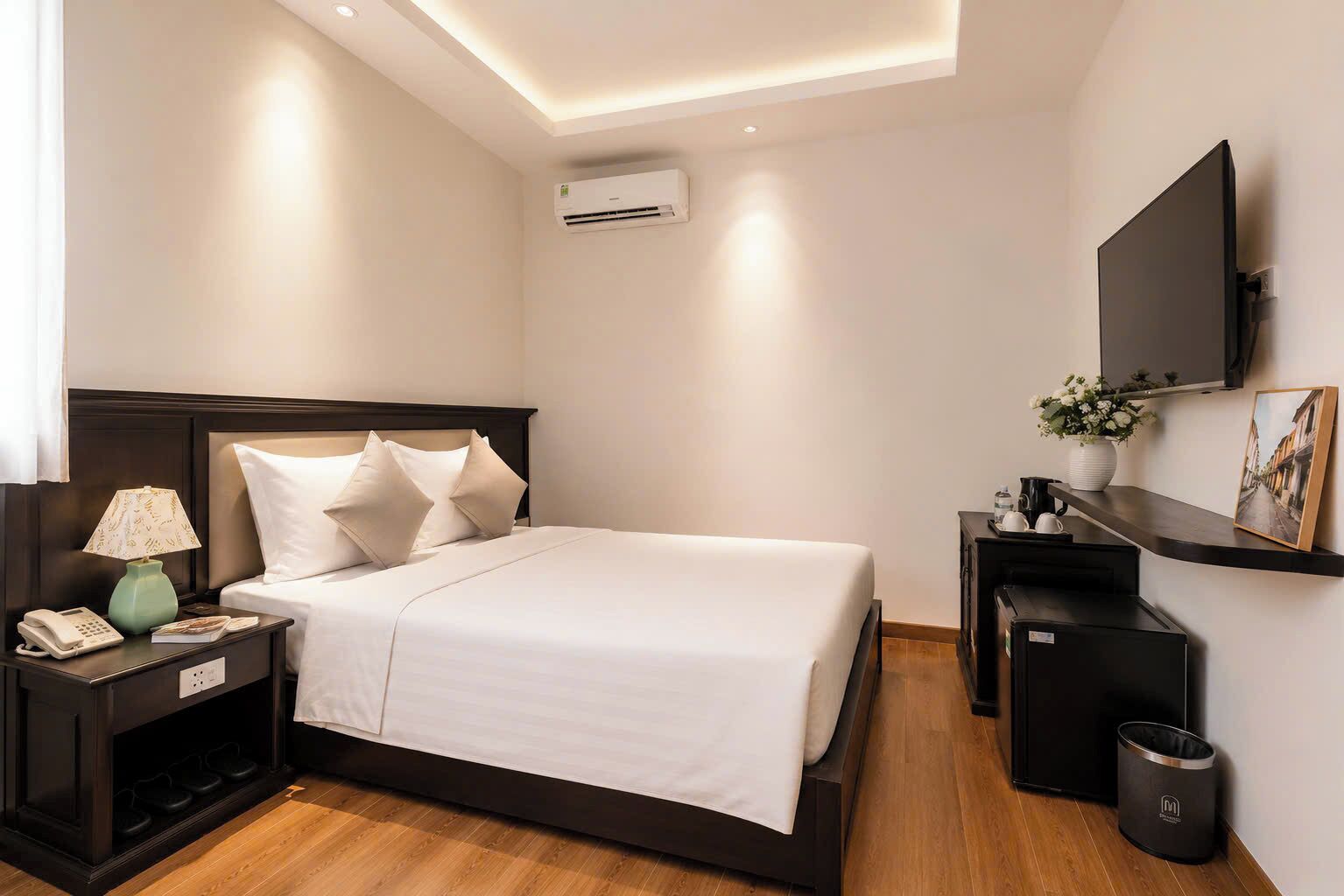 Une chambre d’hôtel comprenant un lit, une table de chevet avec une lampe, un téléviseur, une petite étagère avec un vase et un tableau, ainsi qu’un appareil de climatisation.