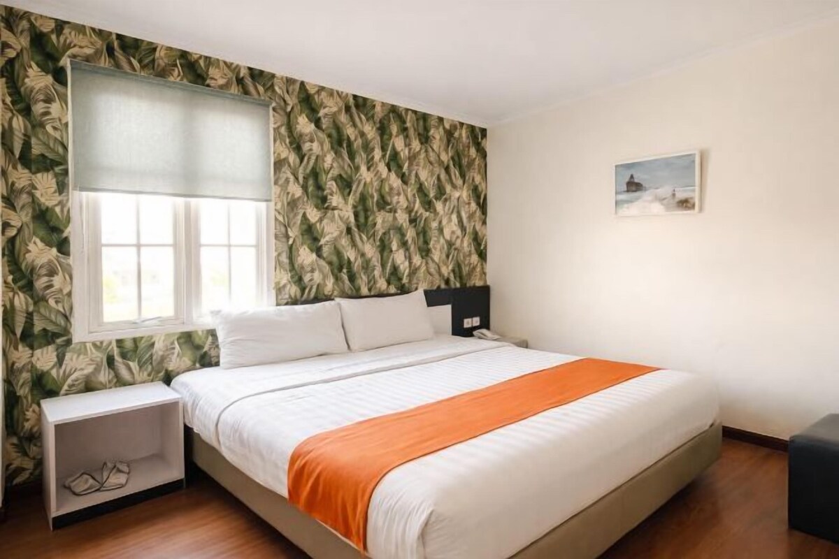 A hotel room with a bed, a nightstand, a window with blinds, a painting on the wall, and a wooden floor.