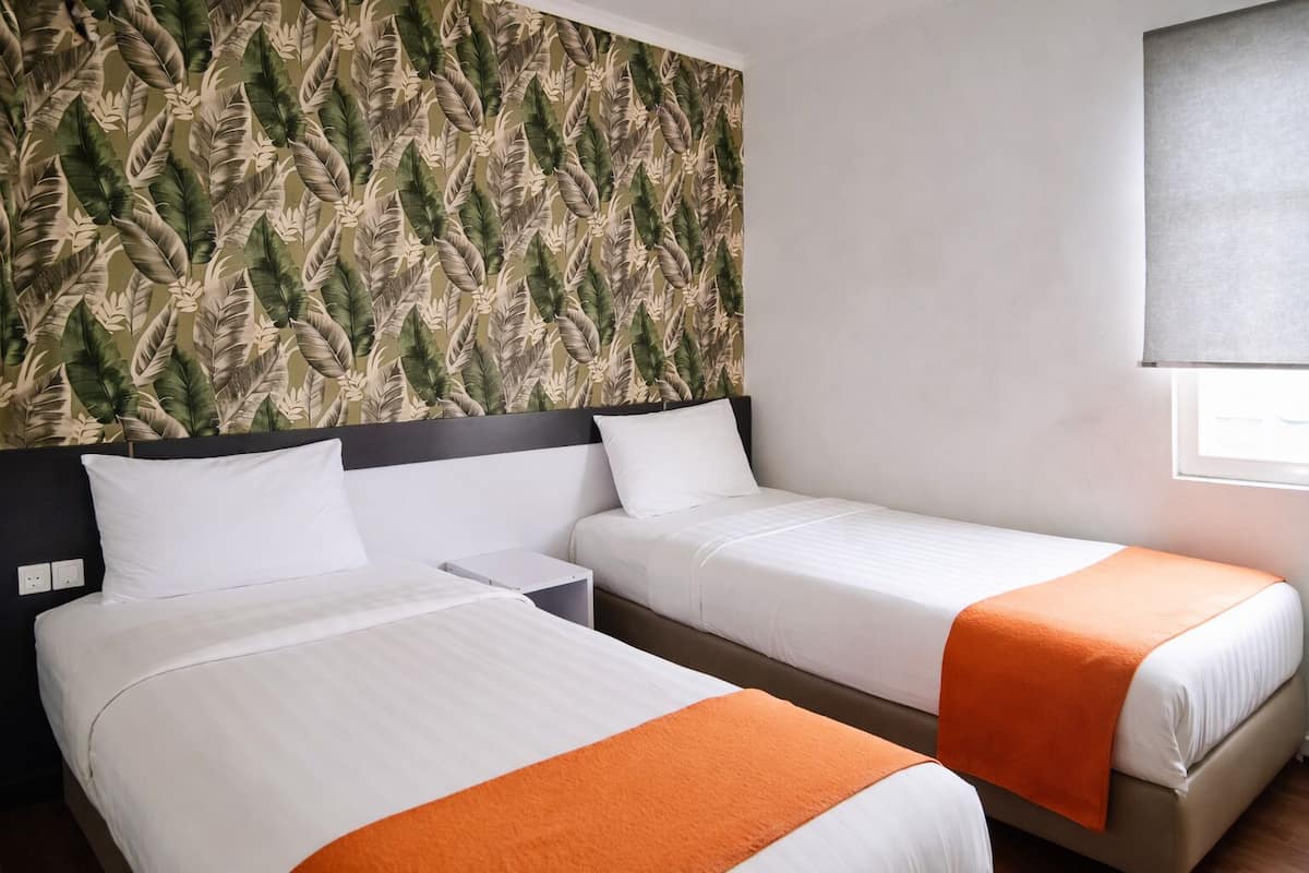 A hotel room with two beds, a headboard with a leaf pattern, white bedding, and an orange throw blanket.