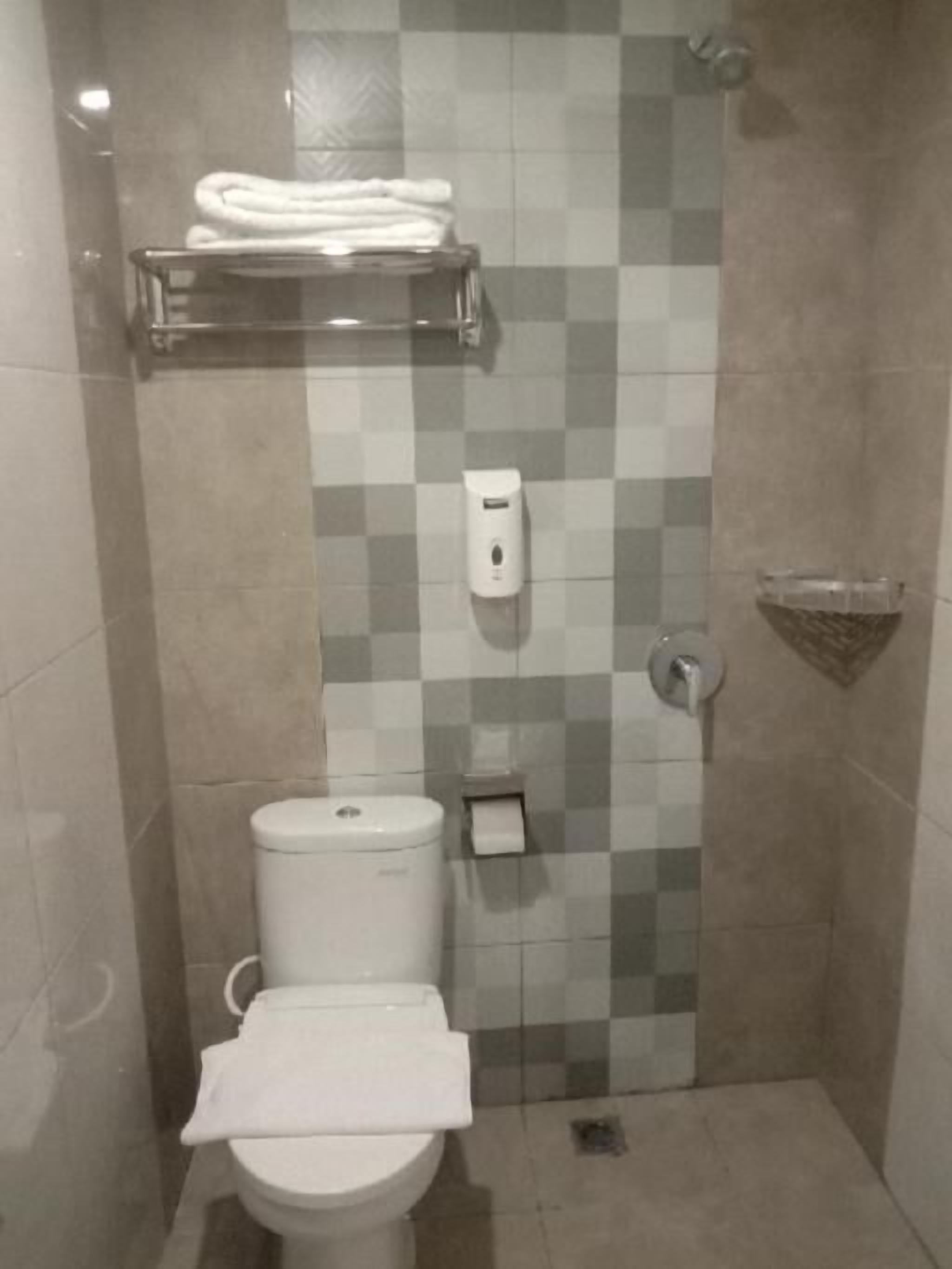 A small, tiled bathroom with a toilet, a towel rack, a soap dispenser, and a shower area.