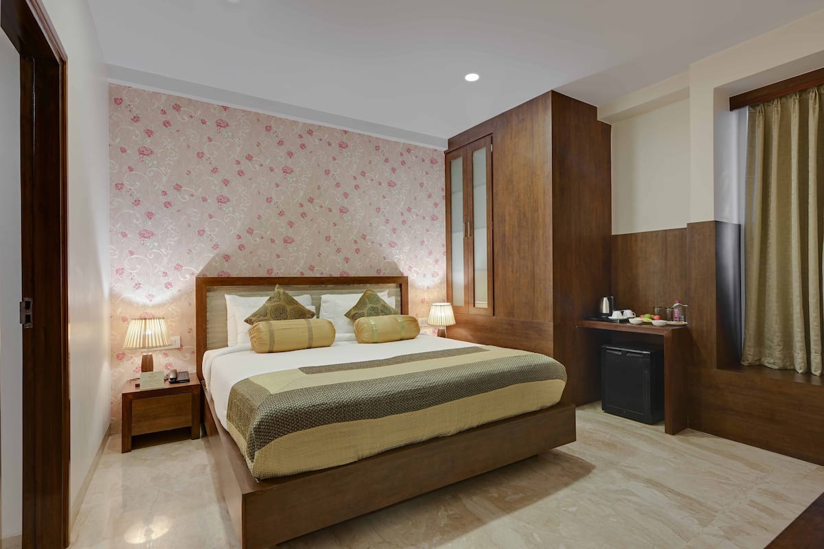 Een slaapkamer met een groot bed, een nachtkastje, een kaptafel, een dressoir, een spiegel, een raam met gordijnen en een klein kitchenette-gedeelte.