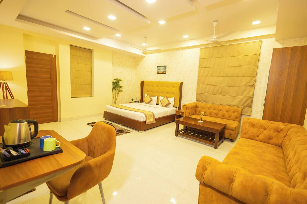 A hotel room with a bed, a sofa, a coffee table, a desk with a kettle, and a chair.
