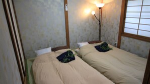 A room with two beds, each with a beige bedspread and a dark blue pillow. There is a wall-mounted light fixture on the right side of the image.