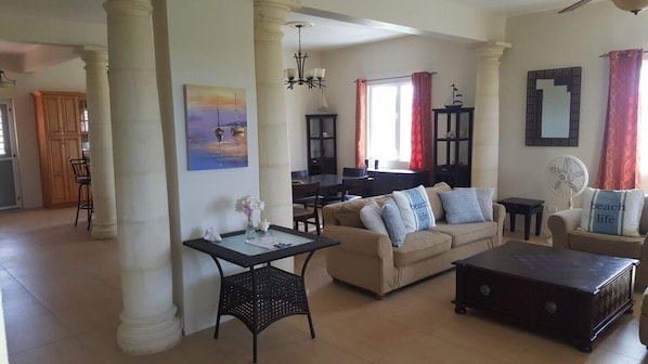 A living room with a beige sofa, a coffee table, a dining area with a table and chairs, and a kitchen in the background.