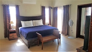 Een slaapkamer met een bed, nachtkastje, stoel, ventilator en gordijnen.