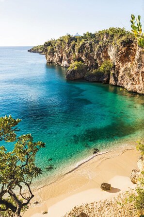 A coastal landscape with clear turquoise waters, sandy beach, rocky cliffs, and sparse vegetation.