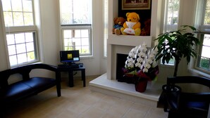 A living room with a fireplace, a desk with a computer, a large teddy bear, and a potted plant.