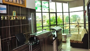 A reception area with a reception desk, a sofa, and a large window offering a view of buildings and trees. - KPA Grand Palace (Sriperumbudur)