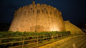 Een historisch stenen kasteel met een grote centrale toren en een omliggende muur, verlicht in de nacht.