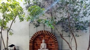 A person meditating on a purple mat in a garden with a statue and brick archway in the background. - Kelaniya Heritage (Kiribathgoda)