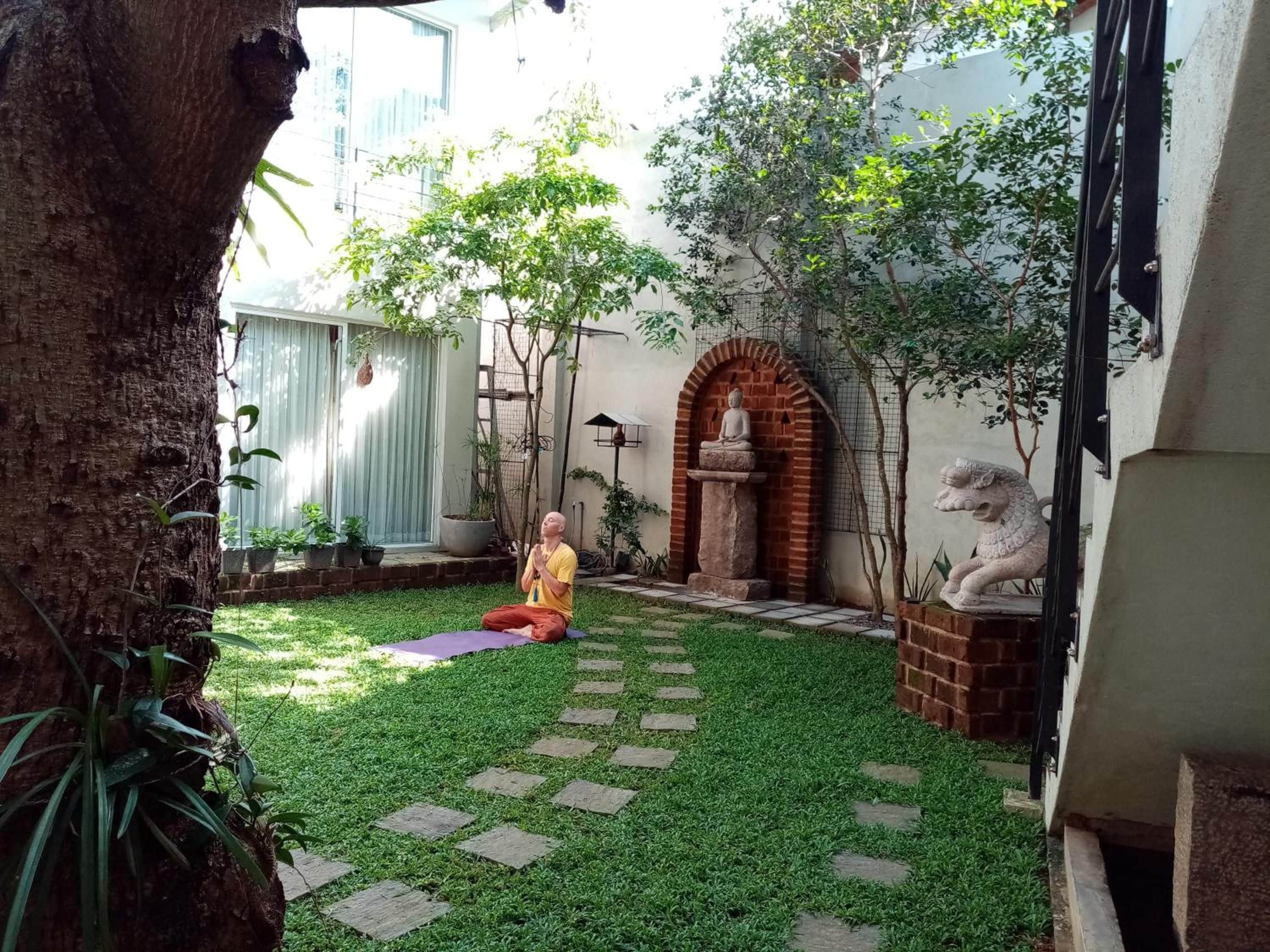 A courtyard with a person meditating on a mat, surrounded by greenery and a statue.