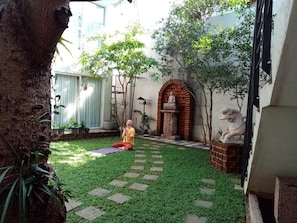 A courtyard with a person meditating on a mat, surrounded by greenery and a statue.