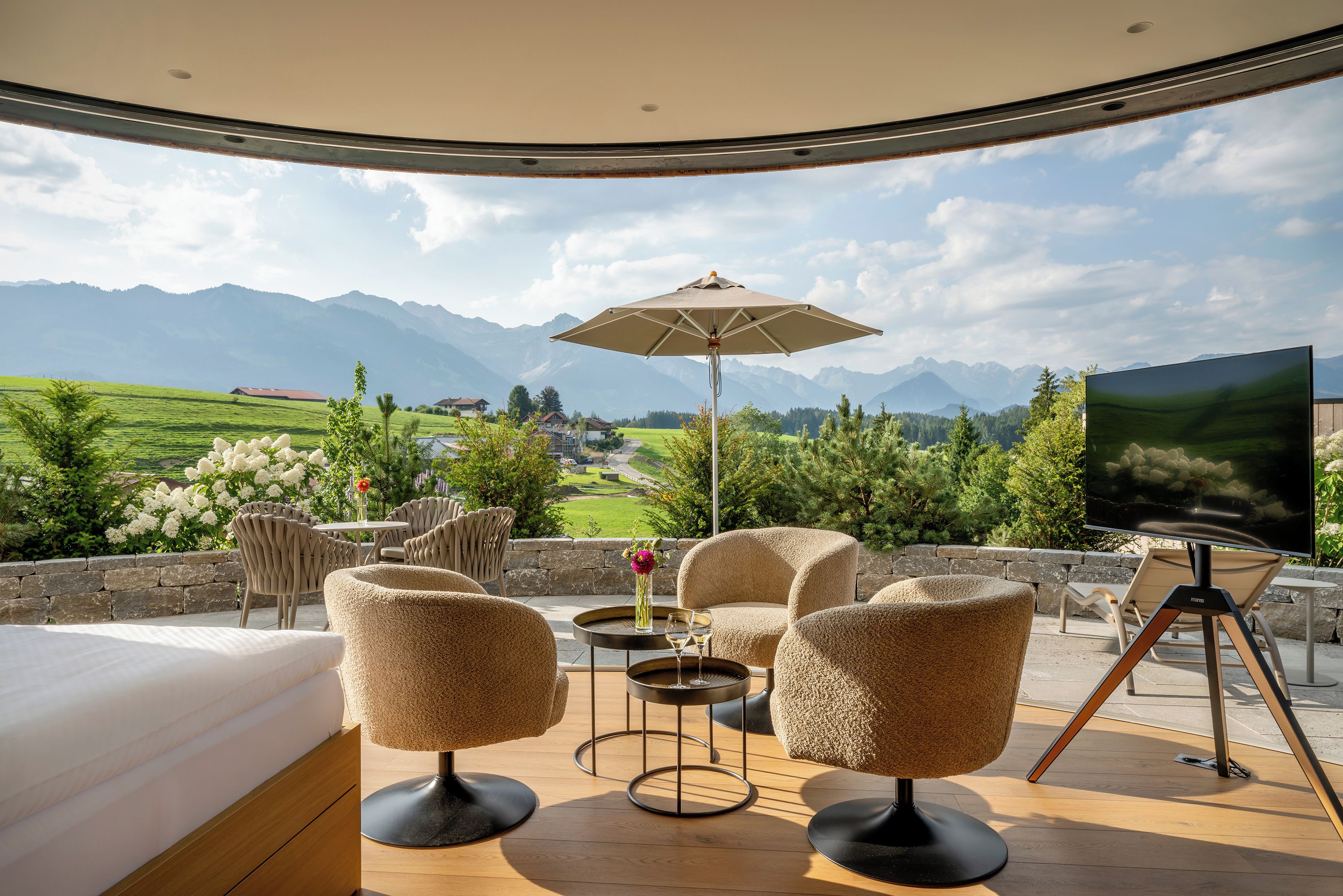 Ein sonniger Außenbereich mit Sitzgelegenheit und Blick auf Berge, ein Tisch mit Blumenvase und ein Fernseher.