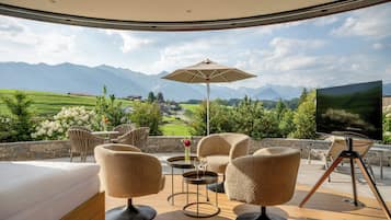 Ein sonniger Außenbereich mit Sitzgelegenheit und Blick auf Berge, ein Tisch mit Blumenvase und ein Fernseher.