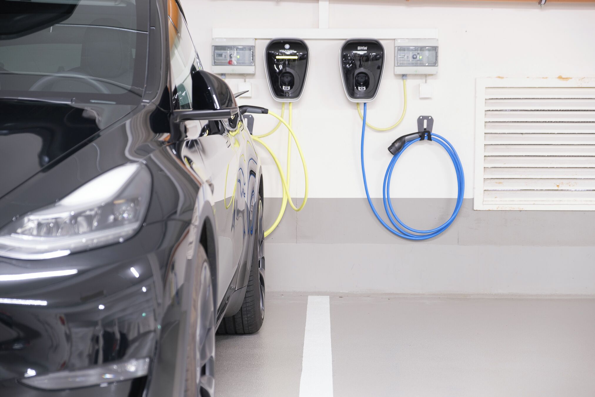 Ein Auto wird an einer Ladestation mit zwei Anschlüssen, einem gelben und einem blauen, aufgeladen. Im Hintergrund ist eine weiße Wand mit einem Lüftungsgitter zu sehen.