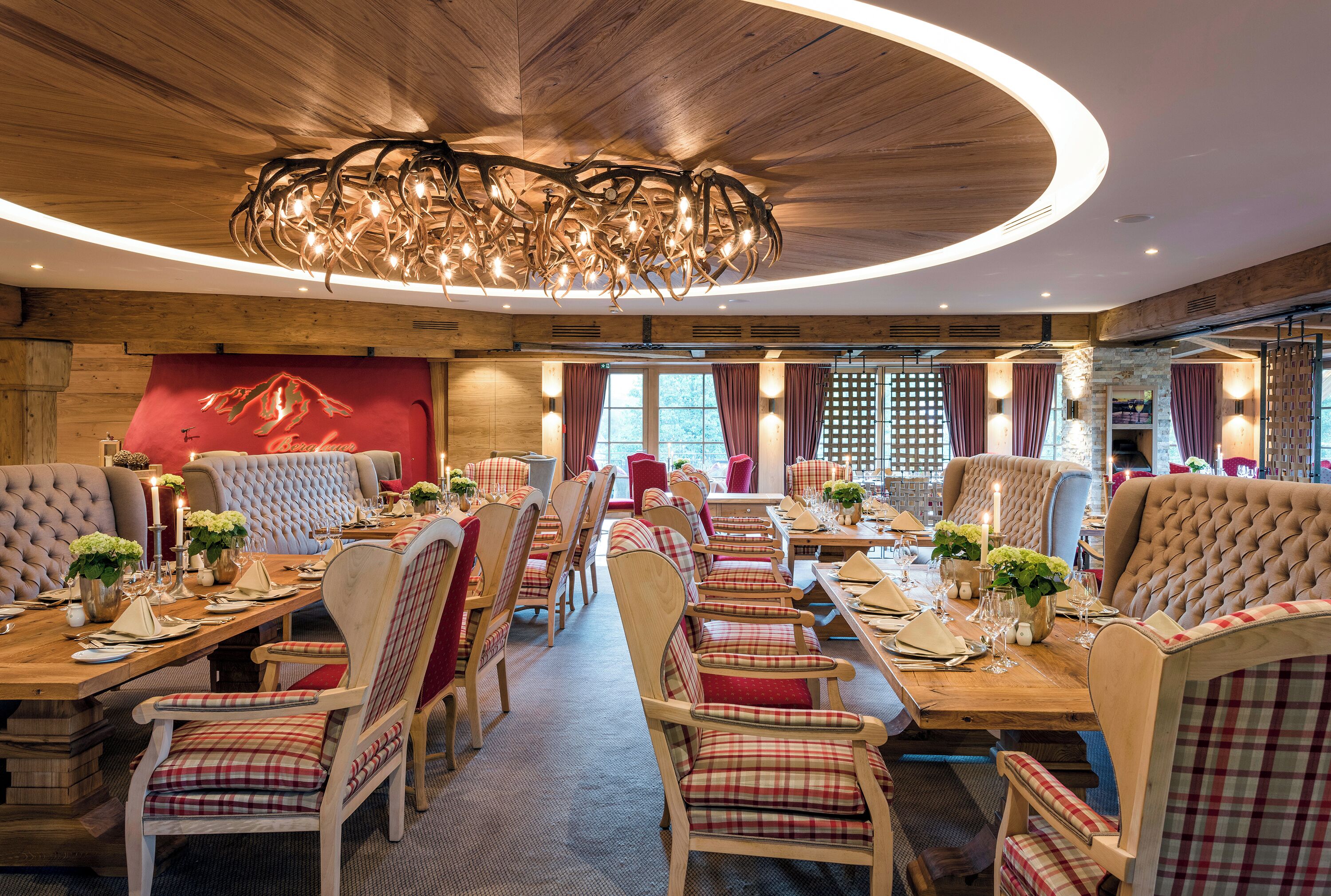 Ein Restaurant mit Holzbalkendecke und einer Kristallleuchter. Die Tische sind mit rot-weiß karierten Tischdecken gedeckt, und es stehen Blumentöpfe darauf.