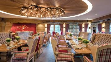 Ein Restaurant mit Holzbalkendecke und einer Kristallleuchter. Die Tische sind mit rot-weiß karierten Tischdecken gedeckt, und es stehen Blumentöpfe darauf.