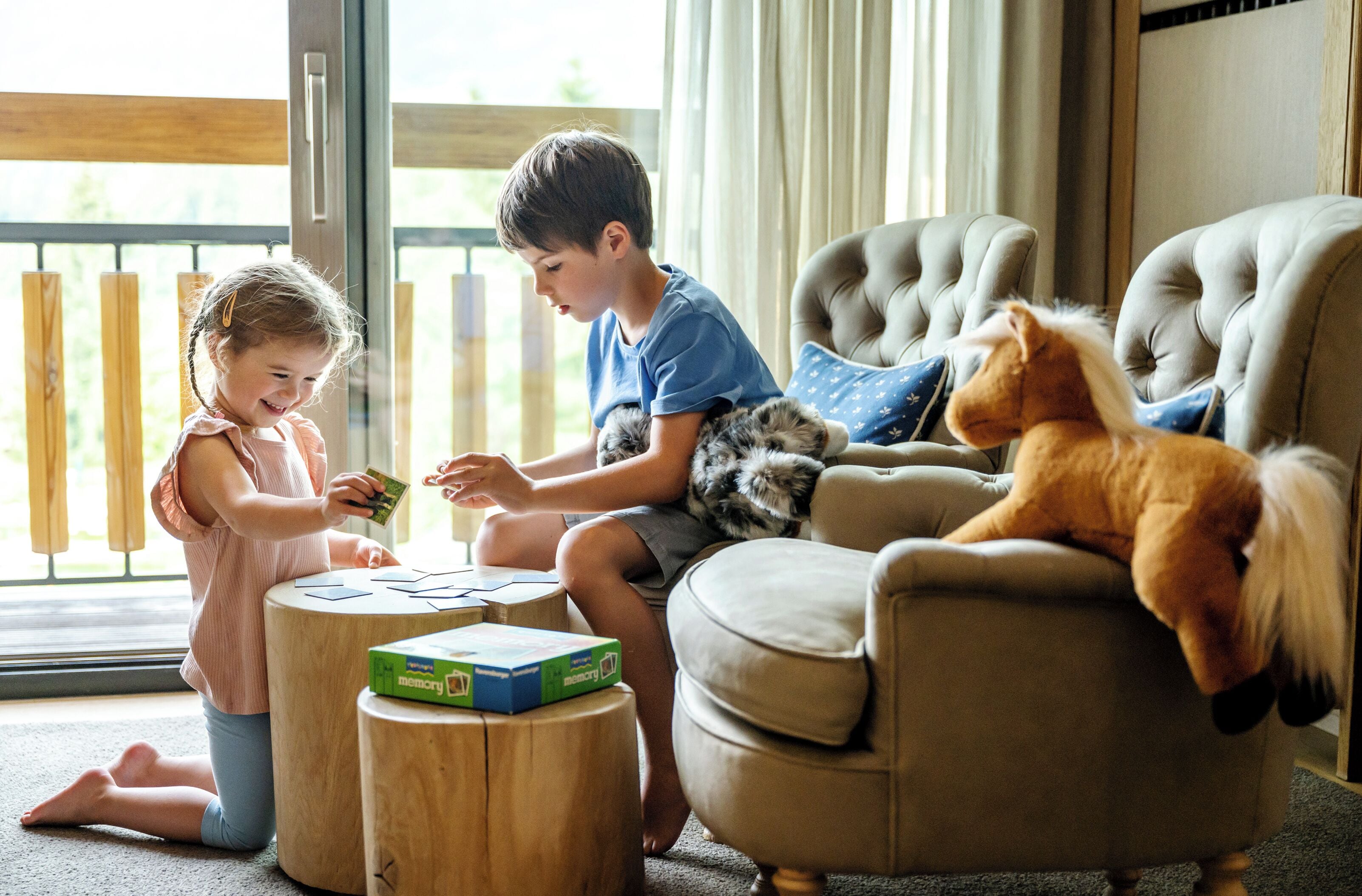 Two children playing with a toy on a wooden stool in a room with a large window and a plush toy on a chair.