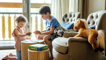 Two children playing with a toy on a wooden stool in a room with a large window and a plush toy on a chair.
