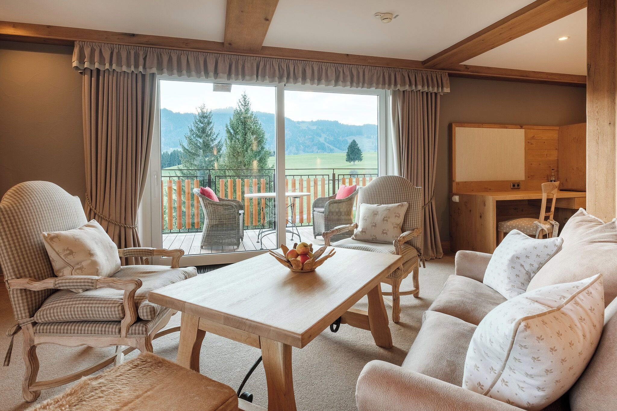 A cozy room with a wooden table, two rocking chairs, a sofa, and a balcony with a view of mountains.