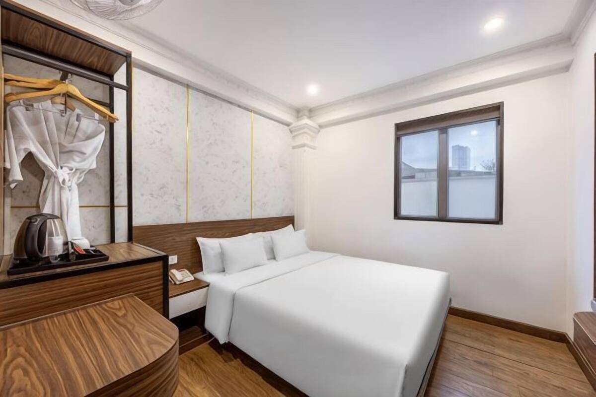 Ein Hotelzimmer mit einem großen Bett, einem Nachttisch aus Holz, einem Spiegel, einem Bademantel und einem Fenster mit Blick auf die Stadt.