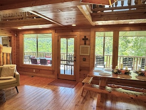 A wooden cabin interior with a cozy seating area, a dining table with candles, and large windows offering a view of the forest.