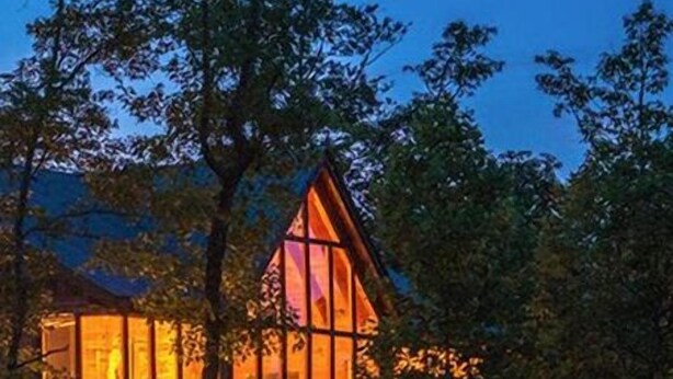 Una baita dal tetto a forma di triangolo, immersa tra gli alberi e illuminata da luci calde dall'interno.