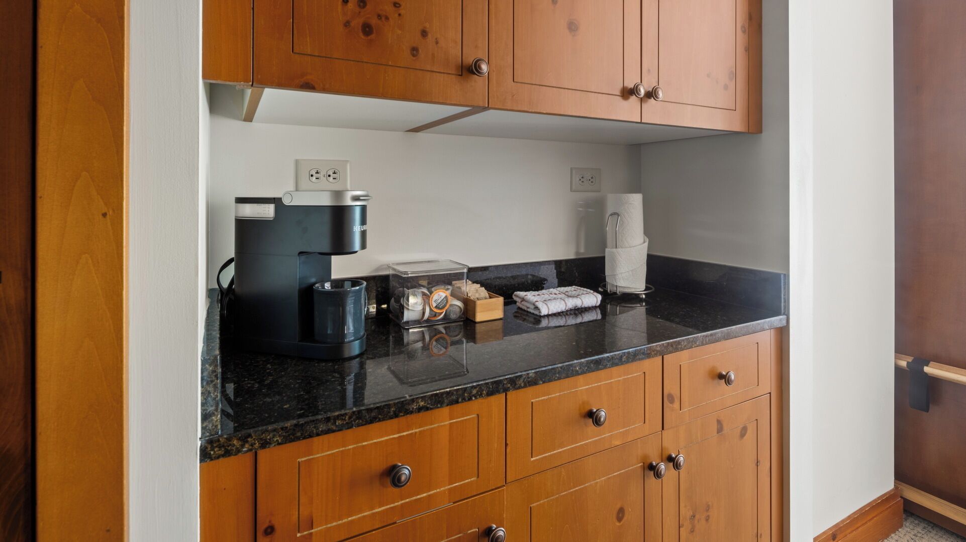 A kitchen with wooden cabinets, a coffee maker, a kettle, and a trash bin.