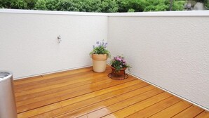 Una terraza de madera con dos macetas, una con flores moradas y otra con flores rosadas, y una pared blanca al fondo.