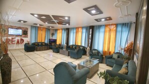 A spacious living room with multiple sofas and a coffee table, a TV mounted on the wall, and a large window with curtains. - Hotel Sheetal Regency (Mathura)