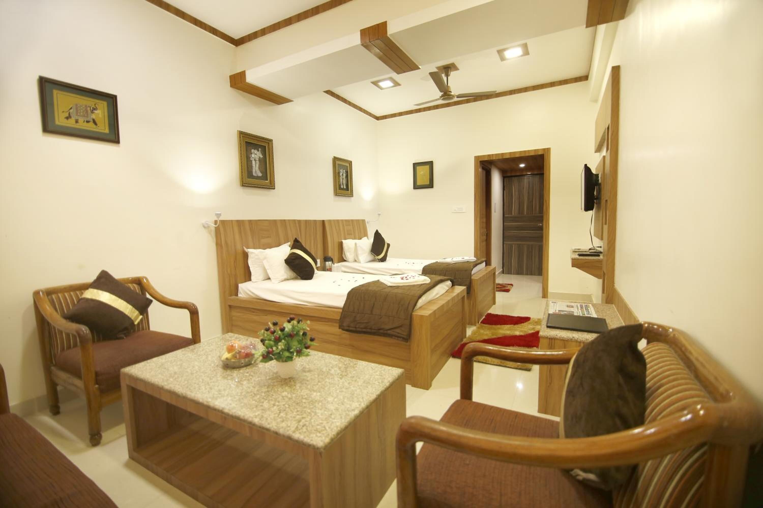 Habitación de hotel con cama, dos sillas de madera, una mesita con un florero y televisor en la pared.