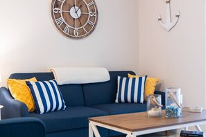 Un salon avec un canapé bleu marine, des coussins à rayures jaunes et bleues, une horloge en bois et une décoration en forme d’ancre au mur.