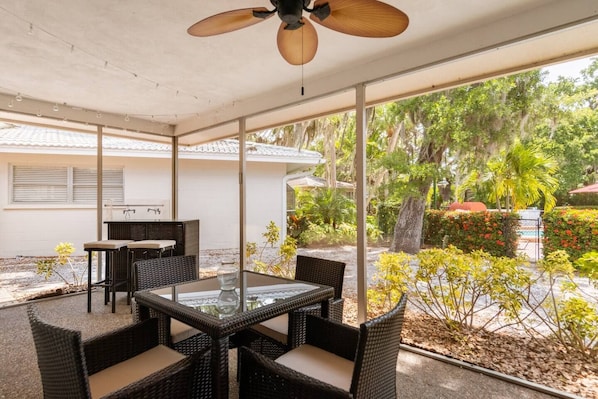 Une terrasse extérieure aménagée avec des meubles en osier, une table à plateau en verre et un ventilateur de plafond.