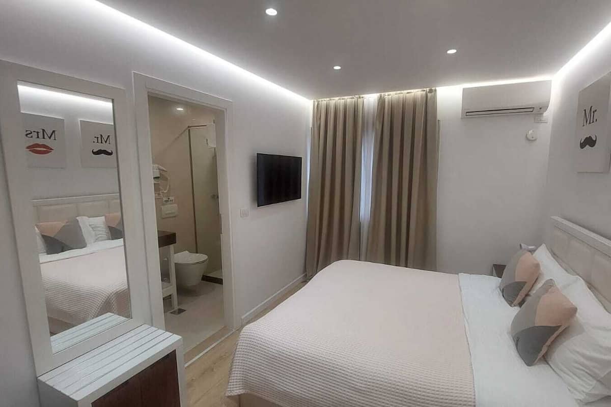 Habitación de hotel con cama, espejo, televisión y baño visible a través de una puerta abierta.