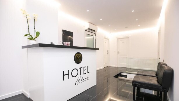 A hotel reception area with a white reception desk, a black bench, and a glass partition. - Hotel Elior  (Tirana)