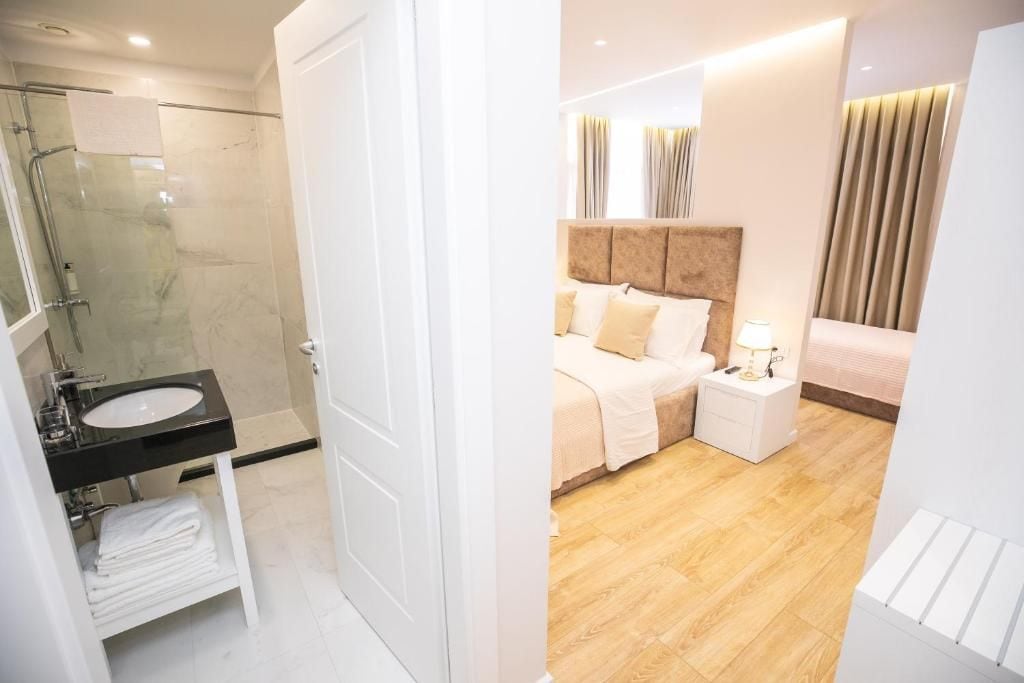 A modern hotel room with a glass shower enclosure, a white sink, a neatly made bed, and a bedside table with a lamp.