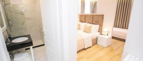 A modern hotel room with a glass shower enclosure, a white sink, a neatly made bed, and a bedside table with a lamp.
