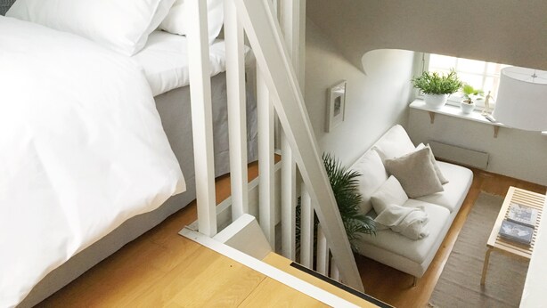 A modern bedroom with a bed, a sofa, a staircase, and a window with plants. - Roost Pursimiehenkatu (Helsinki)