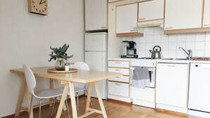A kitchen with white cabinets, a wooden table, two chairs, a clock on the wall, a coffee maker, and a kettle. - Roost Pursimiehenkatu (Helsinki)