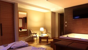 Una camera d'albergo con un letto grande, un'area salotto con una sedia e un tavolo, una TV a muro e una vasca da bagno separata.