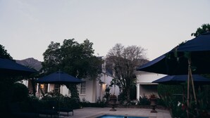 A pool area with umbrellas, seating, and a building with a garden. - Les Chambres (Franschhoek)