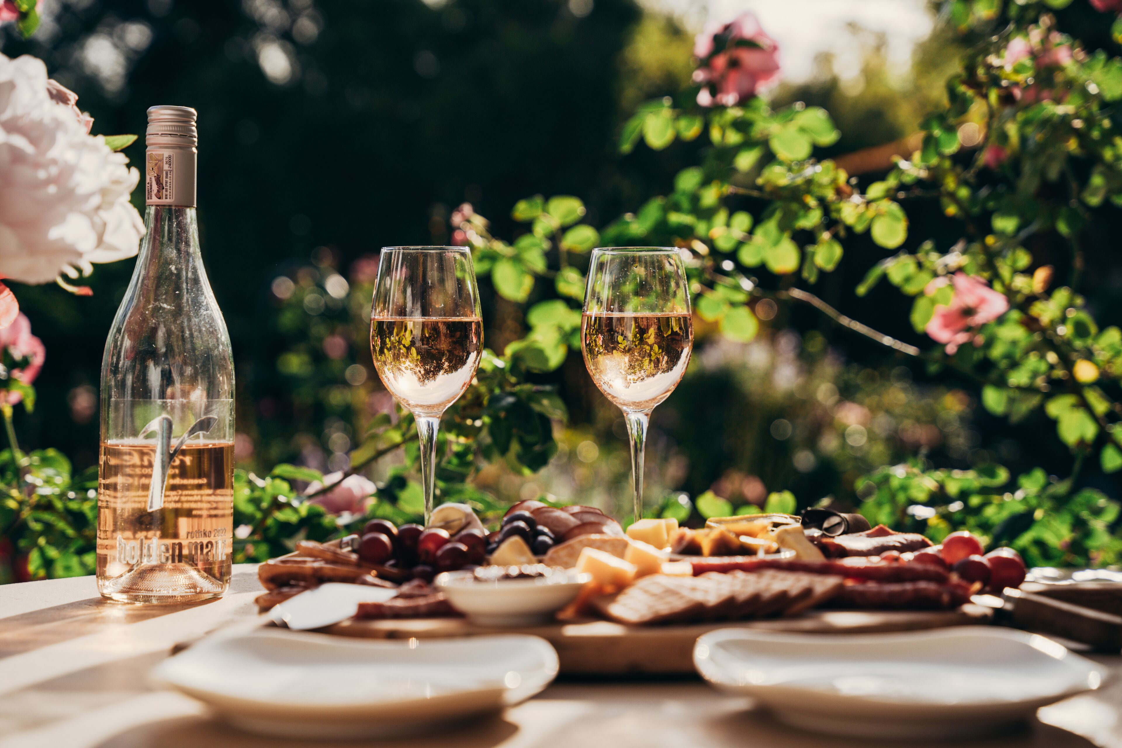 A table with a bottle of wine, two glasses, and a variety of cheeses and fruits.
