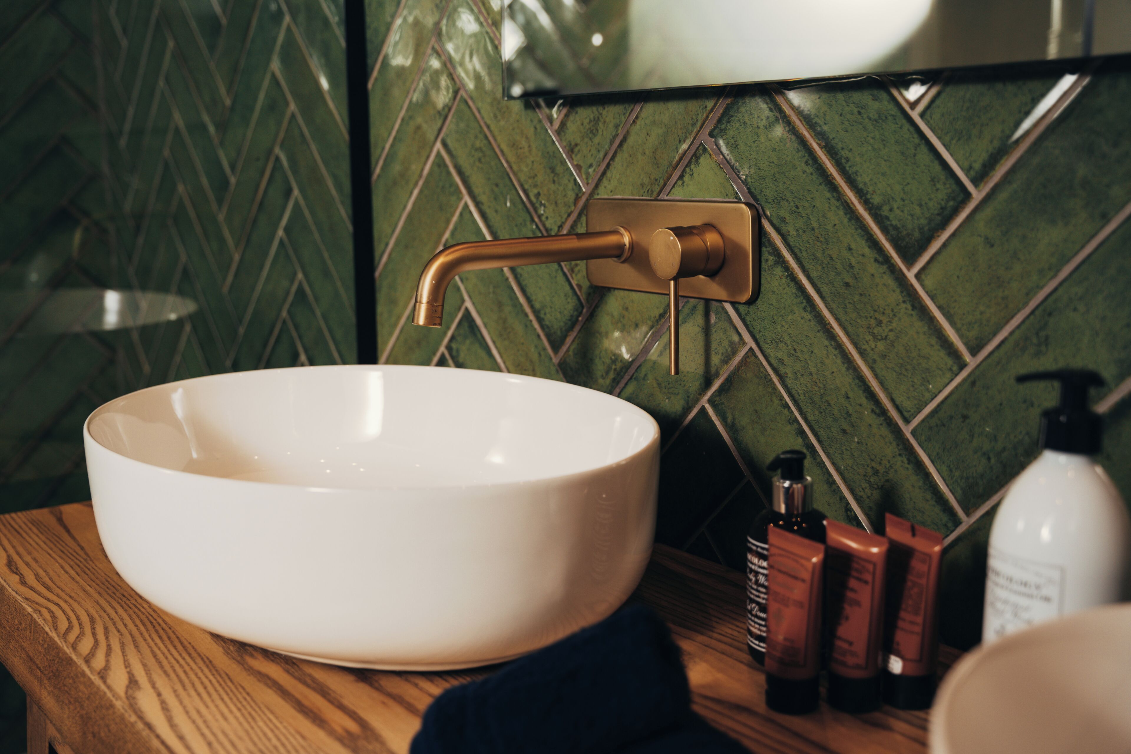 A bathroom with a white vessel sink, a gold faucet, and a green herringbone tile backsplash.