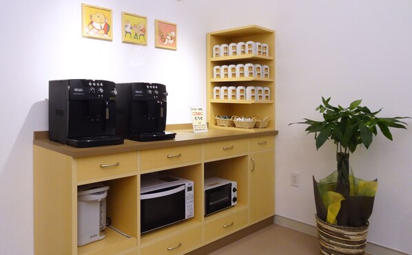 A kitchenette with two coffee makers, a microwave, a small refrigerator, and a shelf with jars. There are three framed pictures on the wall and a potted plant.
