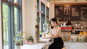 Uma mulher sentada a uma mesa num café, com um quadro de menu ao fundo.
