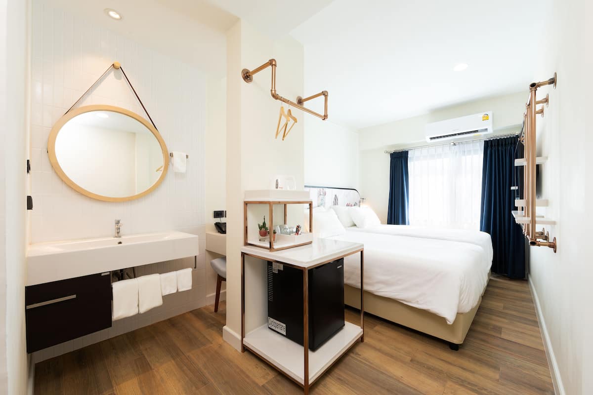 Una habitación de hotel moderna con una cama blanca, un espejo redondo, una mesita con una planta y un equipo de aire acondicionado instalado.
