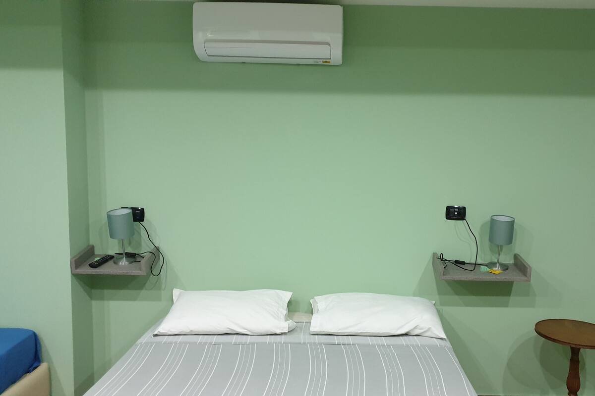 A room with a bed, two bedside tables, a small round table, and an air conditioning unit on the wall.